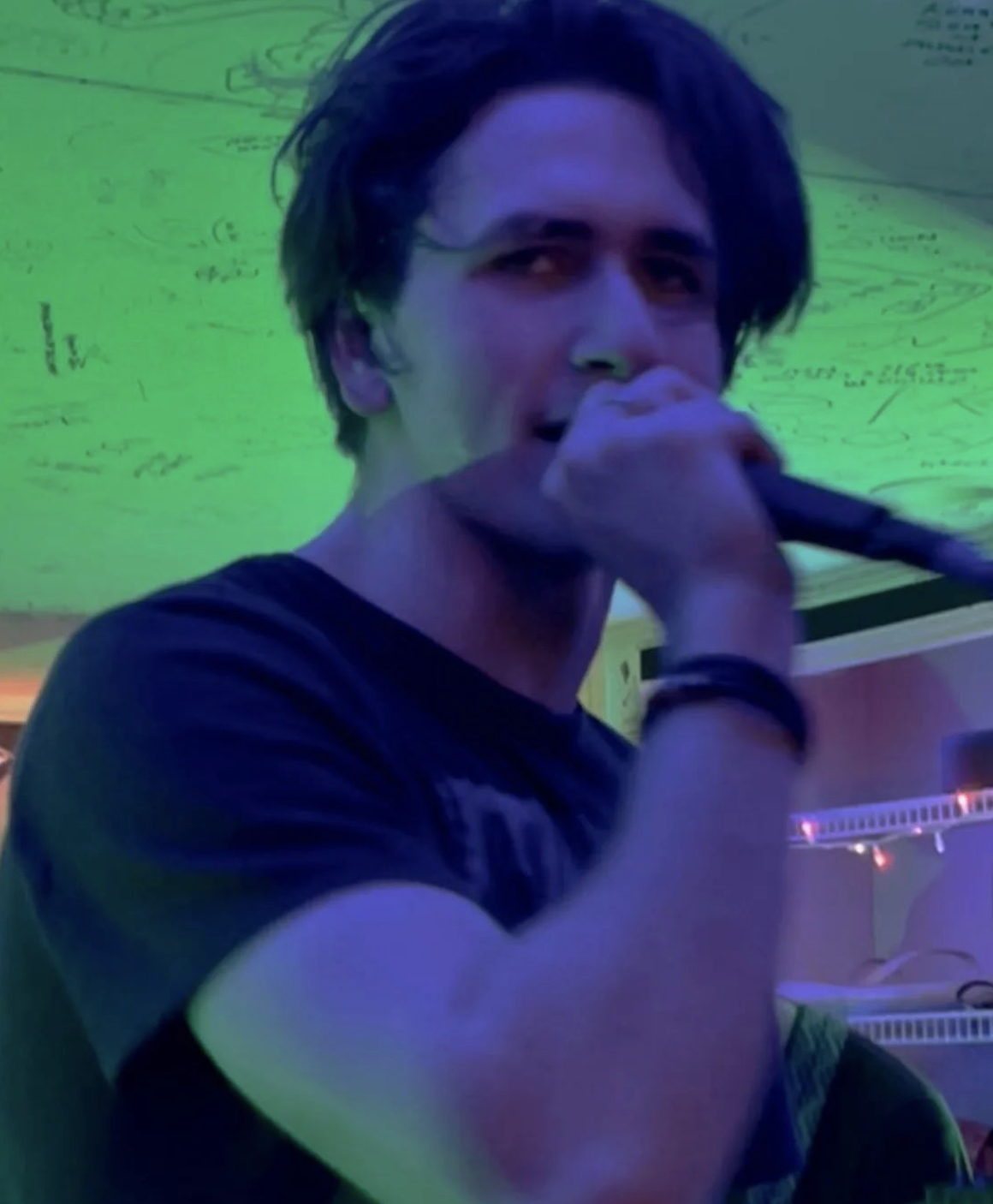 A singer holding a microphone mid-performance, lit by green and blue lighting, with a casual indoor setting in the background.